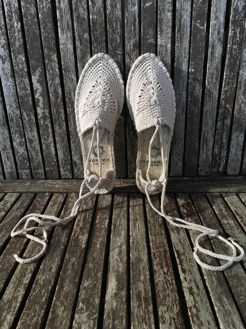 K&ouml;nnte beinhalten: Ein Paar beige geh&auml;kelte Espadrilles-Sandalen mit spitzer Zehenpartie und gewebtem Design. Die Sandalen haben lange, cremefarbene Schn&uuml;rsenkel und werden auf einer verwitterten Holzoberfl&auml;che pr&auml;sentiert. Die Sohlen haben ein gedrucktes Logo.