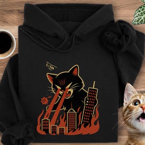 May include: Black hooded sweatshirt with a cartoon cat graphic. The cat is shown with red laser beams destroying buildings. The design includes a helicopter and flames, creating a humorous, action-packed theme. The sweatshirt has a hood and long sleeves.