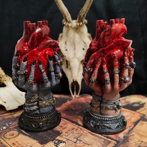 Può includere: Due sculture decorative di mani che tengono un cuore rosso. Le mani sono adornate con anelli e braccialetti. Le sculture sono su una superficie marrone scuro.
