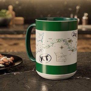 May include: A ceramic coffee mug with a map design. The mug features a white body with green accents on the handle, rim, and base. The map shows a route with the text "KT" and the number "240".