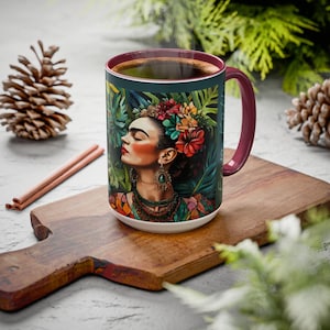 May include: A white ceramic mug with a burgundy rim and handle. The mug features a colorful illustration of Frida Kahlo with flowers in her hair, surrounded by tropical foliage.