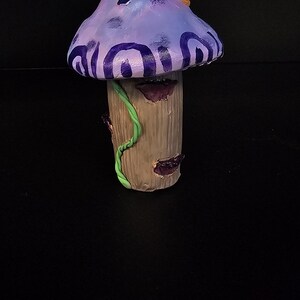 May include: A purple and brown mushroom figurine with a green vine wrapped around the stem. The mushroom cap has a swirl pattern and a small blue square.