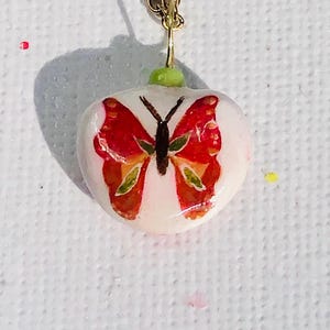 May include: A white heart-shaped pendant necklace with a red and orange butterfly design. The necklace has a gold chain.