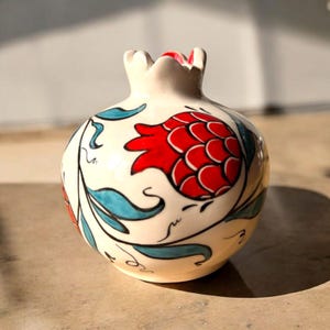 Może przedstawiać: Biały ceramiczny wazon w kształcie granatu z czerwono-niebieskim wzorem kwiatowym. Wazon jest ręcznie malowany z misternymi detalami.