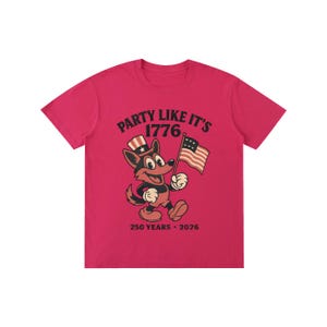May include: A bright pink t-shirt featuring a cartoon fox holding an American flag. The fox wears a patriotic hat and the shirt reads "PARTY LIKE IT'S 1776" with "250 YEARS &bull; 2026" below.