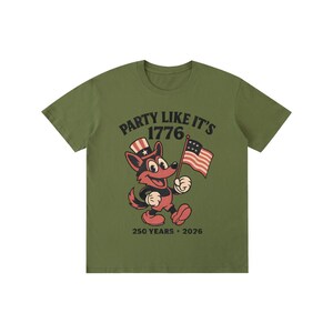 May include: Olive green t-shirt with the text "PARTY LIKE IT'S 1776" in black and a cartoon fox wearing a patriotic hat and holding an American flag. The shirt also includes "250 YEARS &bull; 2026" in black.