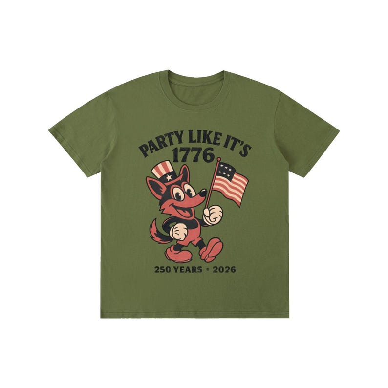 May include: Olive green t-shirt with the text "PARTY LIKE IT'S 1776" in black and a cartoon fox wearing a patriotic hat and holding an American flag. The shirt also includes "250 YEARS &bull; 2026" in black.