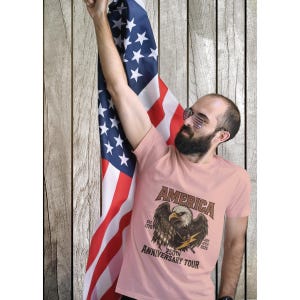 Puede incluir: Camiseta rosa claro con la palabra "AMERICA" sobre un águila. La camiseta también tiene el texto "EST. 1776" y "250TH ANNIVERSARY TOUR JULY 4TH 2026". Una bandera americana está en el fondo.