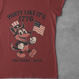May include: Red t-shirt featuring a cartoon fox wearing a patriotic hat and holding an American flag. The shirt has the text "PARTY LIKE IT'S 1776" and "250 YEARS - 2026".
