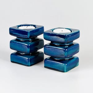 May include: Two matching teal blue ceramic candle holders. Each holder has a square top with a recessed area for a candle, stacked on two additional square sections. The holders have a glossy finish and are set against a white background.
