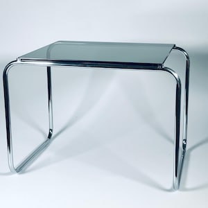 May include: A chrome-plated metal side table with a glass top. The table has a simple, modern design with a rectangular top and four legs.
