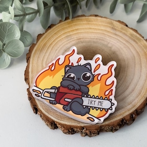 Peut inclure: Un autocollant transparent représentant un chat de dessin animé tenant une tronçonneuse avec des flammes en arrière-plan. La tronçonneuse porte l'inscription "TRY ME" sur la lame. Le chat est gris avec de grands yeux.