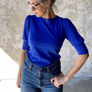 May include: A woman wearing a royal blue short-sleeved top and blue jeans. The top has a round neck and puffed sleeves. The jeans are a classic denim style. The woman is wearing glasses and has her hair up in a bun.