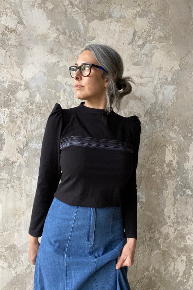 May include: A black long-sleeved shirt with a high neckline and puffed shoulders. The shirt has a thin horizontal stripe design in a lighter shade of gray. The shirt is paired with a blue denim skirt.
