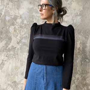 May include: A black long-sleeved shirt with a high neckline and puffed sleeves. The shirt has a thin horizontal stripe design across the chest. The shirt is paired with a blue denim skirt.