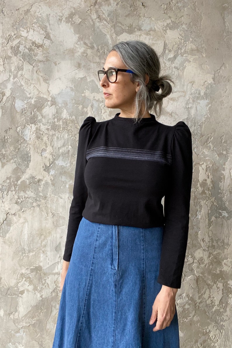 May include: A black long-sleeved shirt with a high neckline and puffed sleeves. The shirt has a thin horizontal stripe design across the chest. The shirt is paired with a blue denim skirt.