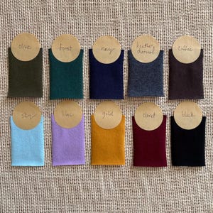 Puede incluir: Diez muestras de tela rectangulares dispuestas en dos filas, con los colores olive, forest, navy, heather charcoal, coffee, sky, lilac, gold, claret y black. Cada muestra tiene una etiqueta circular de papel color canela con el nombre.