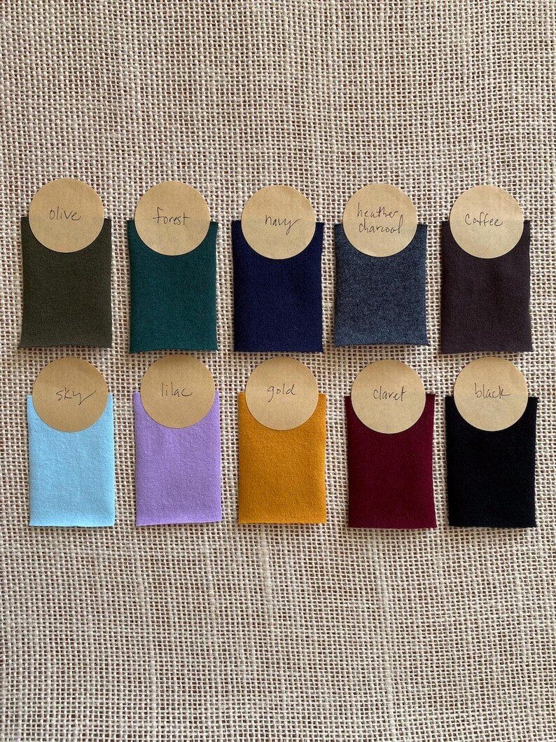 Puede incluir: Diez muestras de tela rectangulares dispuestas en dos filas, con los colores olive, forest, navy, heather charcoal, coffee, sky, lilac, gold, claret y black. Cada muestra tiene una etiqueta circular de papel color canela con el nombre.