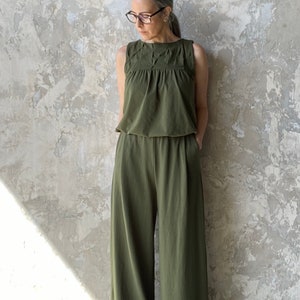 Puede incluir: Un mono sin mangas de color verde oliva con cuello redondo y detalles plisados en los hombros y el pecho. La prenda presenta un cuerpo ajustado que se transforma en un pantal&oacute;n de pernera ancha con una silueta relajada y fluida.