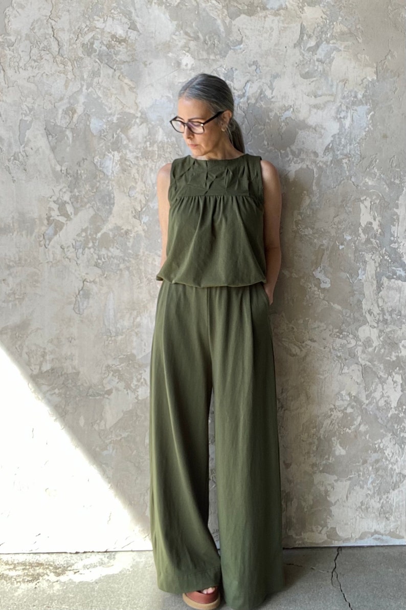 Puede incluir: Un mono sin mangas de color verde oliva con cuello redondo y detalles plisados en los hombros y el pecho. La prenda presenta un cuerpo ajustado que se transforma en un pantal&oacute;n de pernera ancha con una silueta relajada y fluida.