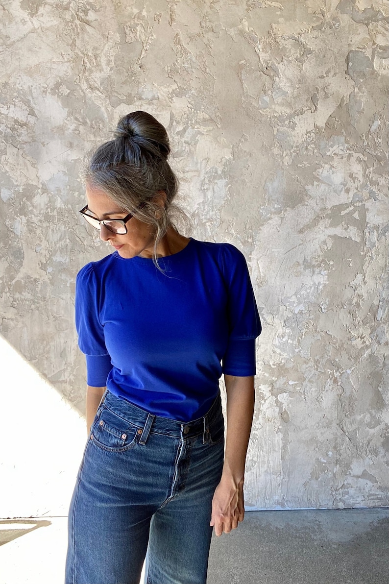 May include: A woman wearing a royal blue short-sleeve top with puffed sleeves and dark wash denim jeans. The top is a solid color, and the jeans have a classic five-pocket design. The background is a textured, neutral-toned wall.