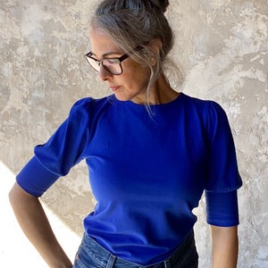 May include: A royal blue short-sleeved top with a round neckline. The top has puffed sleeves and is paired with blue denim jeans. The model is wearing glasses and has their hair up in a bun.