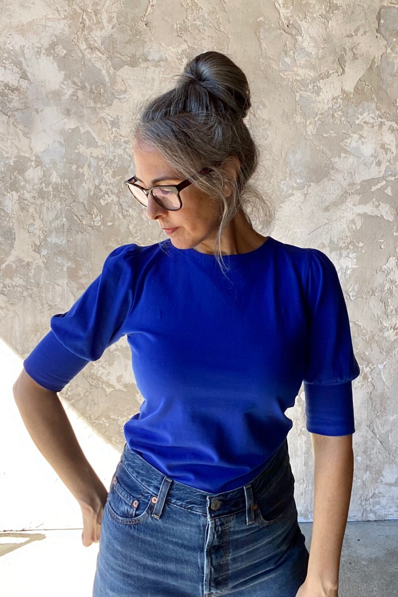 May include: A royal blue short-sleeved top with a round neckline. The top has puffed sleeves and is paired with blue denim jeans. The model is wearing glasses and has their hair up in a bun.
