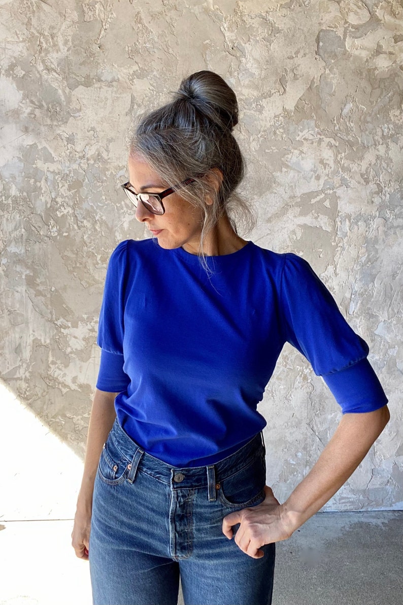 May include: A woman wearing a royal blue short-sleeved top and blue jeans. The top has a round neck and puffed sleeves. The jeans are a classic denim style. The woman is wearing glasses and has her hair up in a bun.
