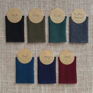 May include: A collection of fabric swatches in various colors, including black, olive, forest green, heather charcoal, teal, navy, and claret. Each swatch is displayed in a small, folded pocket with a circular label indicating the color name.