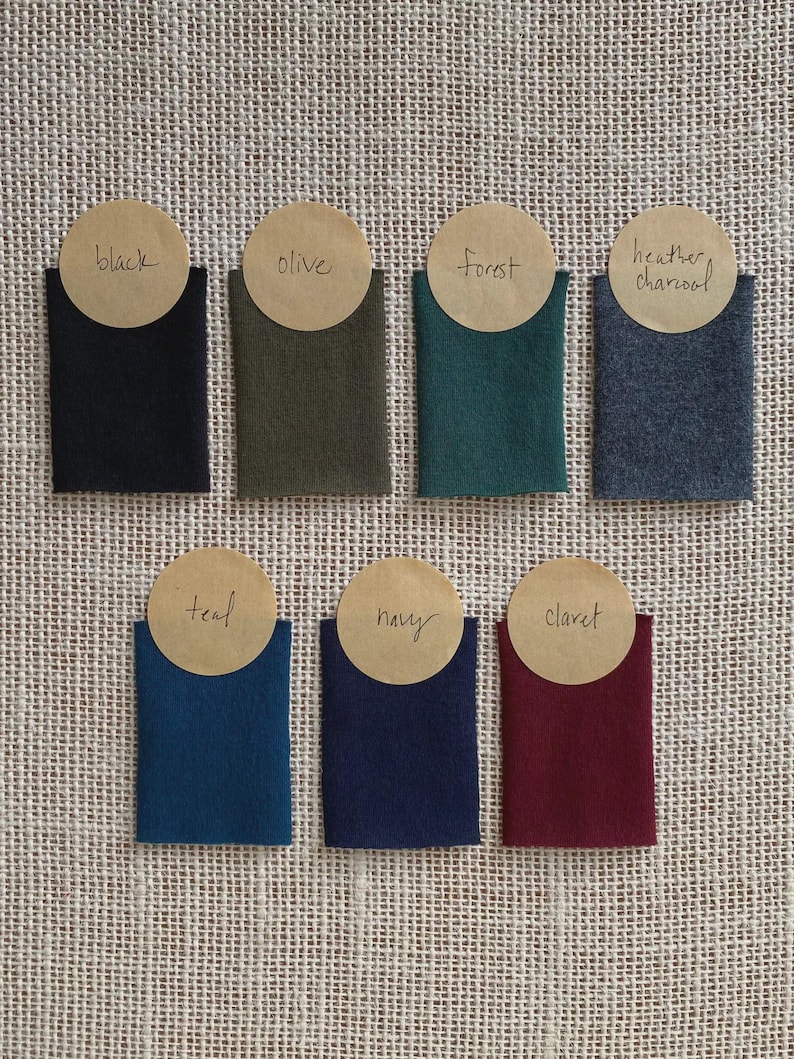 May include: A collection of fabric swatches in various colors, including black, olive, forest green, heather charcoal, teal, navy, and claret. Each swatch is displayed in a small, folded pocket with a circular label indicating the color name.