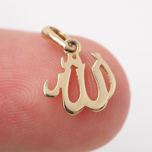 May include: A small, gold-colored pendant in the shape of the Arabic word for "God," Allah. The pendant has a loop at the top for a chain and is displayed against a blurred background.