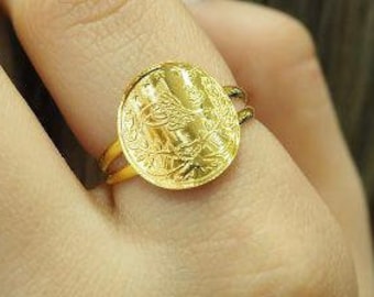 May include: A gold ring with a round, textured design. The ring is worn on the finger of a person's hand.