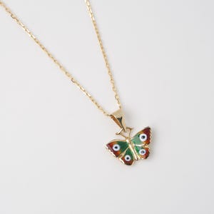 May include: A gold-colored necklace with a butterfly pendant. The butterfly pendant features a green body, red-brown wing tips, and white and blue eye-like details. The necklace is on a white background.