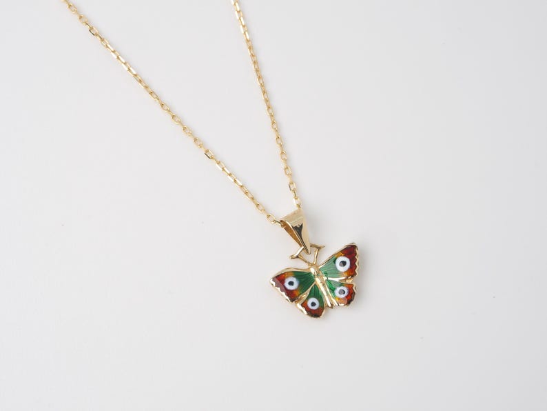 May include: A gold-colored necklace with a butterfly pendant. The butterfly pendant features a green body, red-brown wing tips, and white and blue eye-like details. The necklace is on a white background.