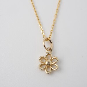 May include: A gold-colored necklace with a daisy pendant. The pendant has six petals and a small, clear gemstone in the center. The chain is delicate and hangs against a white background.