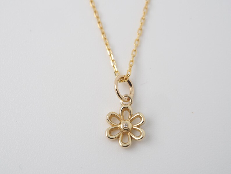 May include: A gold-colored necklace with a daisy pendant. The pendant has six petals and a small, clear gemstone in the center. The chain is delicate and hangs against a white background.
