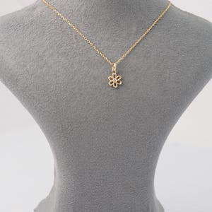 May include: A delicate gold-colored necklace featuring a small, openwork flower pendant. The pendant is suspended from a fine chain, creating a simple and elegant design. The necklace is displayed on a gray mannequin, highlighting the jewelry's details.