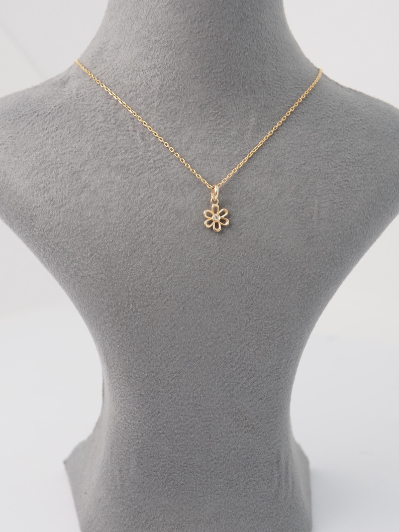 May include: A delicate gold-colored necklace featuring a small, openwork flower pendant. The pendant is suspended from a fine chain, creating a simple and elegant design. The necklace is displayed on a gray mannequin, highlighting the jewelry's details.