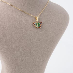 May include: A gold-colored necklace featuring a butterfly pendant. The butterfly pendant is adorned with red, green, and white enamel, and has a delicate chain. The necklace is displayed on a neutral-toned mannequin.