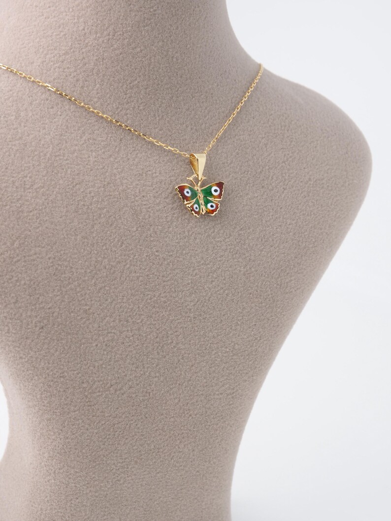 May include: A gold-colored necklace featuring a butterfly pendant. The butterfly pendant is adorned with red, green, and white enamel, and has a delicate chain. The necklace is displayed on a neutral-toned mannequin.