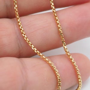 Könnte beinhalten: Eine goldfarbene Box-Kettenhalskette, die aus einer Reihe kleiner, quadratischer Glieder besteht, die in einem durchgehenden, gleichmäßigen Muster verbunden sind. Die Metallkette hat eine polierte, reflektierende Oberfläche, die das Licht einfängt und die komplexe Konstruktion der Glieder zur Geltung bringt.
