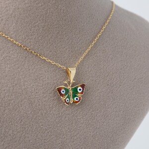 May include: A gold-colored butterfly pendant necklace. The butterfly pendant features red, green, and white enamel detailing. The necklace chain is delicate and gold-colored. The pendant is approximately 1 inch wide.