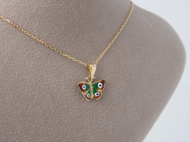 May include: A gold-colored butterfly pendant necklace. The butterfly pendant features red, green, and white enamel detailing. The necklace chain is delicate and gold-colored. The pendant is approximately 1 inch wide.