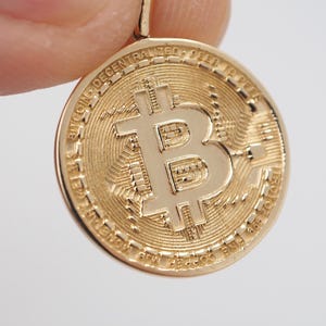 Könnte beinhalten: Ein runder, goldfarbener Anhänger mit einem erhabenen Bitcoin-Symbol in der Mitte. Der Hintergrund der Münze weist ein strukturiertes Leiterplattenmuster auf. Der äußere Rand ist mit dem Text BITCOIN DECENTRALIZED PEER TO PEER versehen.