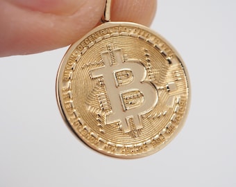 Puede incluir: Un colgante circular de tono dorado con un símbolo de Bitcoin en relieve en el centro. El fondo de la moneda tiene un patrón de circuito impreso texturizado. El borde exterior está grabado con el texto BITCOIN DECENTRALIZED PEER TO PEER.