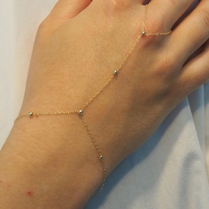 May include: A delicate gold hand chain bracelet with small beaded accents. The bracelet features a fine chain that connects to a ring and wraps around the wrist, creating a minimalist and elegant look. The jewelry is designed to be worn on the hand.