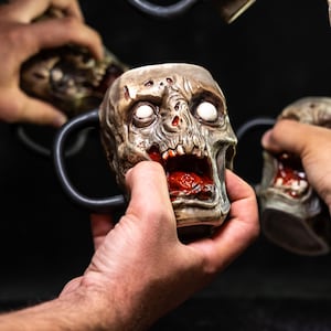May include: Four ceramic zombie head mugs with black handles. The mugs are painted to look like zombie faces with open mouths, showing teeth and blood. The mugs are held in hands.