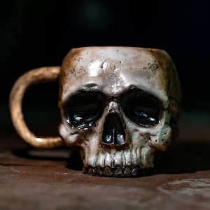 May include: A ceramic skull mug with a white glaze and a detailed skull design. The mug has a handle and is perfect for serving coffee, tea, or other beverages.