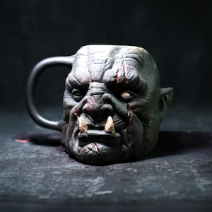 Könnte beinhalten: Eine Keramiktasse in Form eines Ork-Kopfes. Die Tasse ist grau und hat eine detaillierte Skulptur eines Ork-Gesichts mit einer großen Nase, scharfen Zähnen und einer Narbe auf der Stirn.