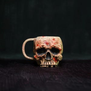 May include: A beige skull-shaped mug with a handle, featuring red splatter details. The mug has a detailed skull design with visible teeth and eye sockets. The mug is set against a dark background.